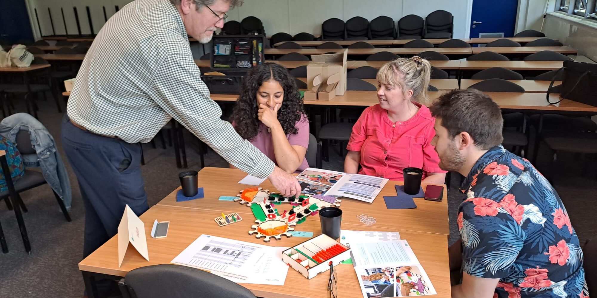 TP_AAPS_Bath_0726 Three adults sitting at a table, playing a brightly coloured game. With a fourth adult leaning over and demonstrating something.